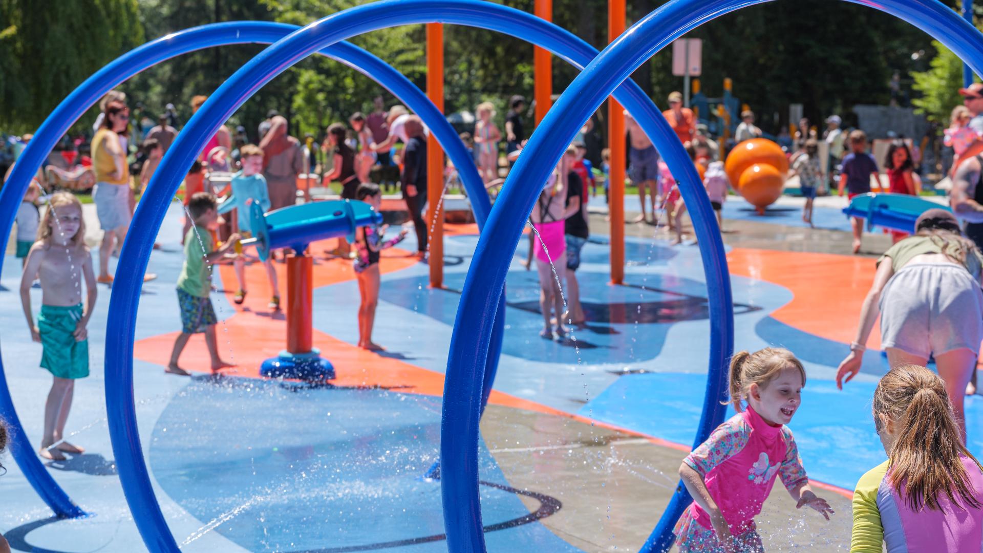 Blue circular spray loops in a spray park with a water canon in the background and kids playing